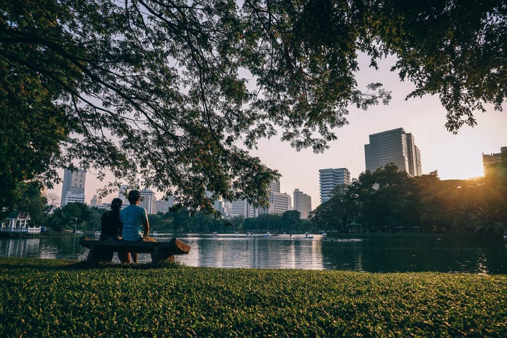 Bangkok am Tag. Fotografiert an einem See. Davor sitzen 2 Menschen auf einer Parkbank.