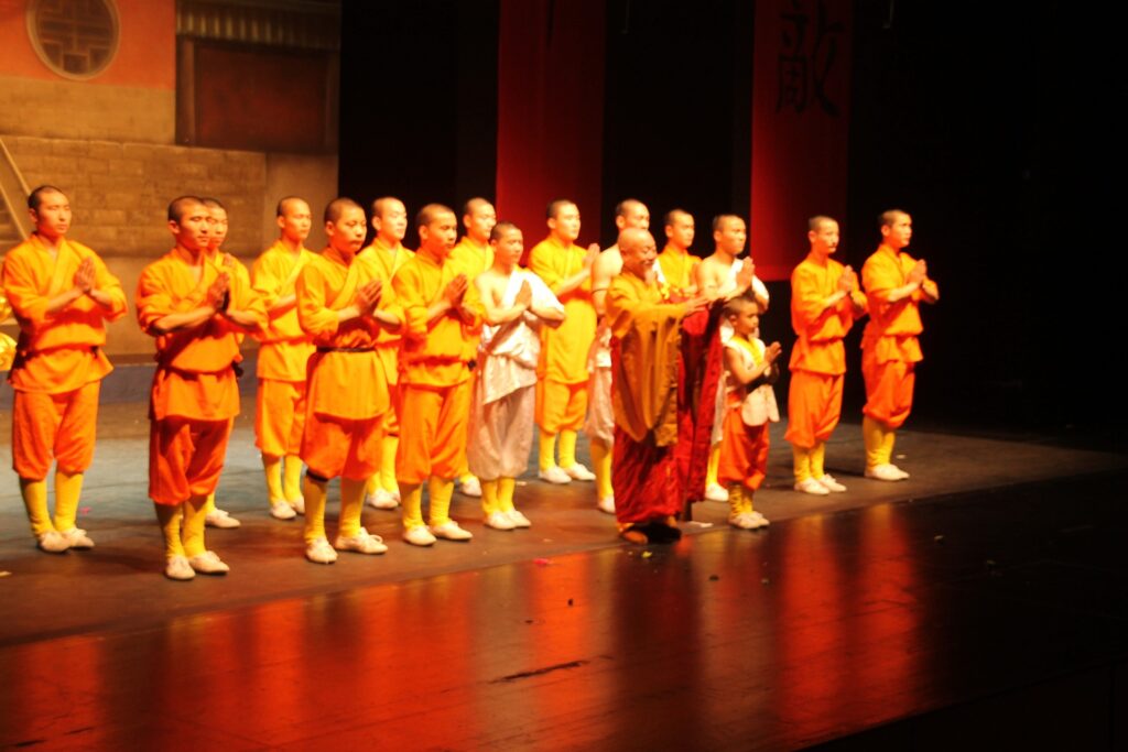 Das Bild zeigt eine Gruppe Shaolin Kämpfer (Mönche), die auf einer Bühne stehen. Vorne steht ein alter Shaolin und neben ihm ein Shaolin Kind. Dahinter steht die Gruppe der Shaolin Kämpfer. Gemeinsam widmen sie sich ihrem Publikum.